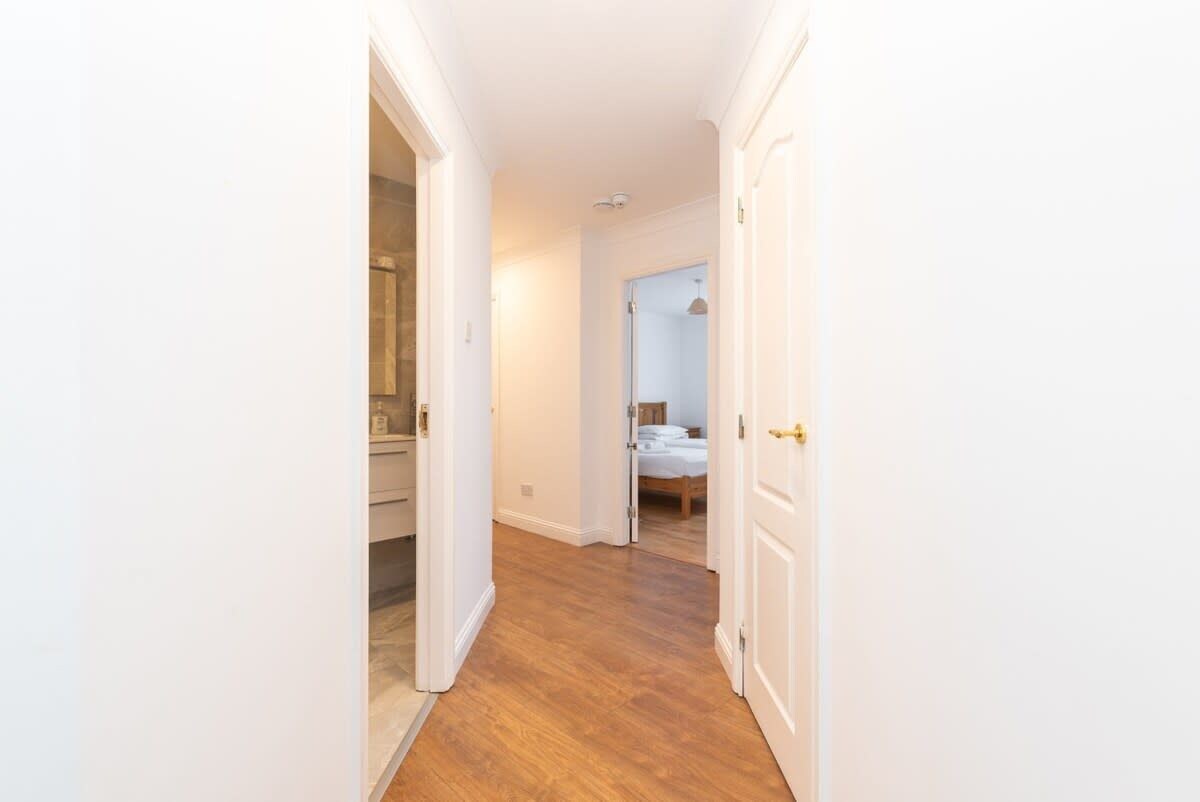 A narrow hallway with wooden flooring leading to a bedroom with a bed and a bathroom with a sink and mirror.