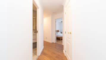 A narrow hallway with wooden flooring leading to a bedroom with a bed and a bathroom with a sink and mirror.
