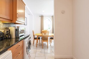 A compact kitchen with a washing machine, a dining table with chairs, and a view through a window. - Ferncourt Stepaside S (Dublin)