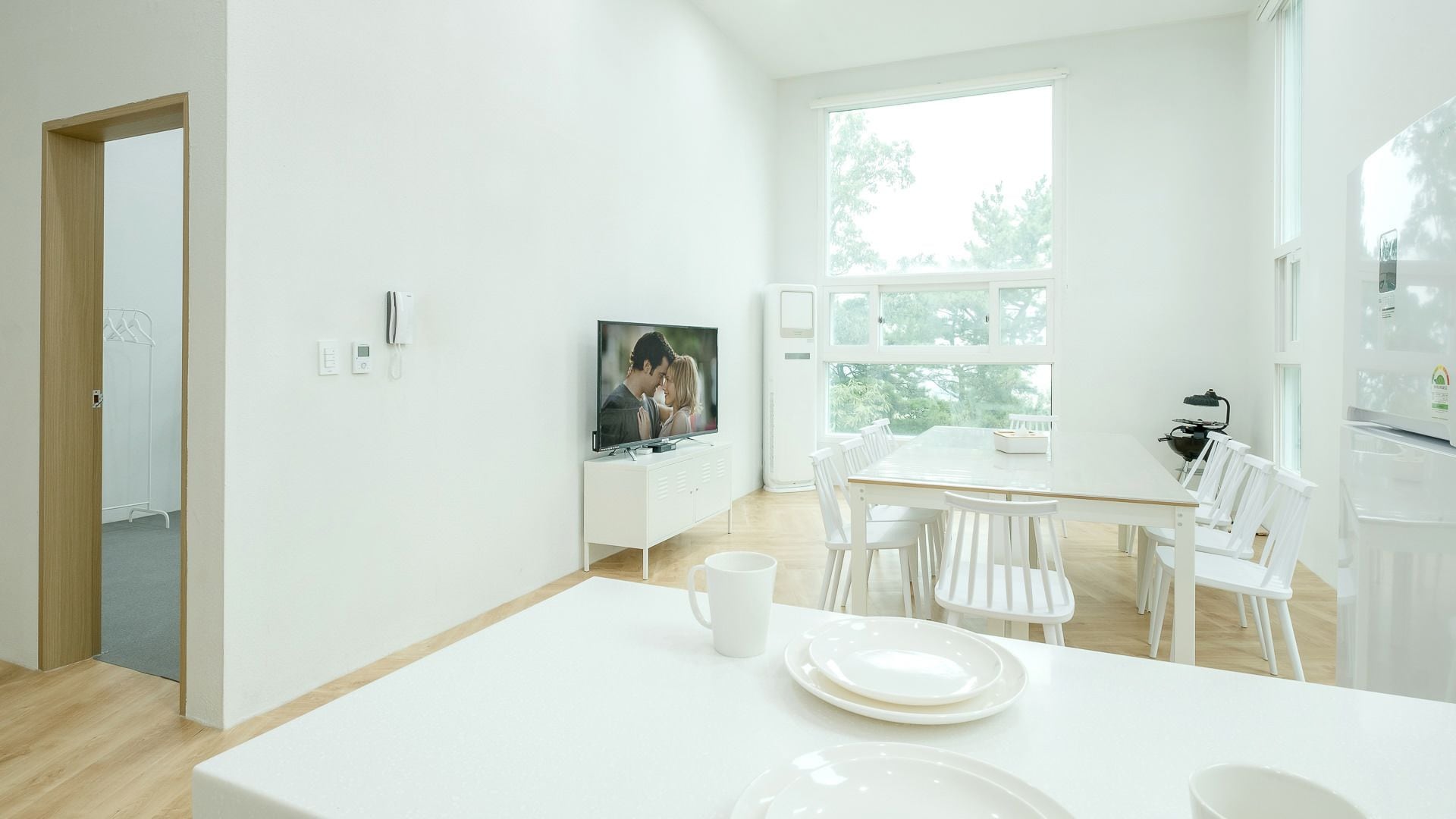 Uma sala de estar moderna, em tons de branco, com área de jantar, televisão e vista para o verde lá fora.