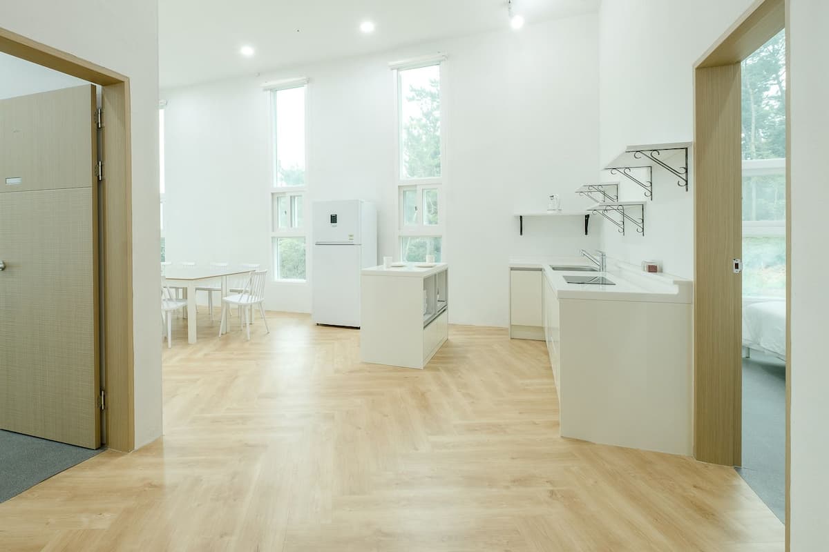 A modern, well-lit room with a wooden floor, a kitchenette, a dining area with chairs, and large windows providing natural light.