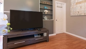 Ein modernes Wohnzimmer mit einem Flachbildfernseher auf einem Holzboden, einer weißen Tür und einem dekorativen Regal mit Gegenständen, darunter ein Fisch und eine Muschel.