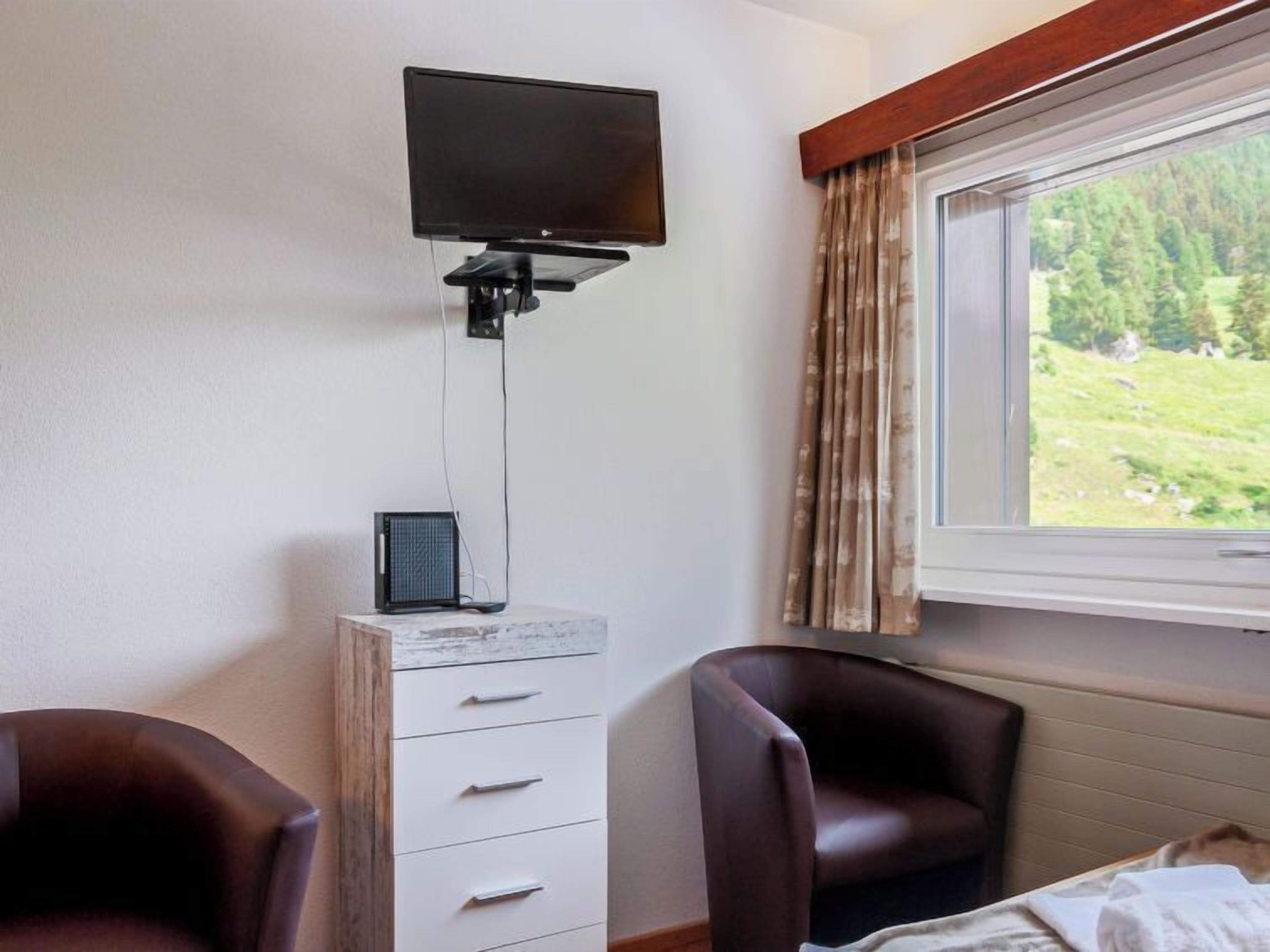 A room with a bed, a desk with a flat-screen TV, a chair, and a window with a view of greenery.