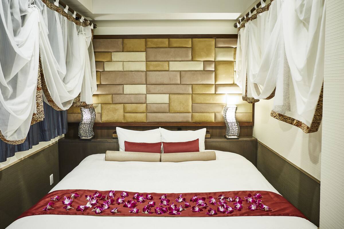 A neatly made bed with a red floral bedspread, a headboard with a textured panel design, and two bedside lamps with decorative vases.