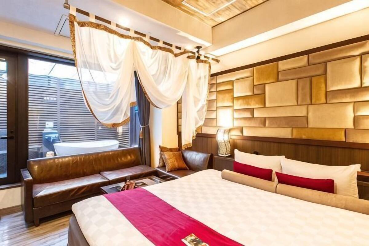 A bedroom with a large bed, a bedside table, a nightstand, a window with curtains, and a view of the outside.