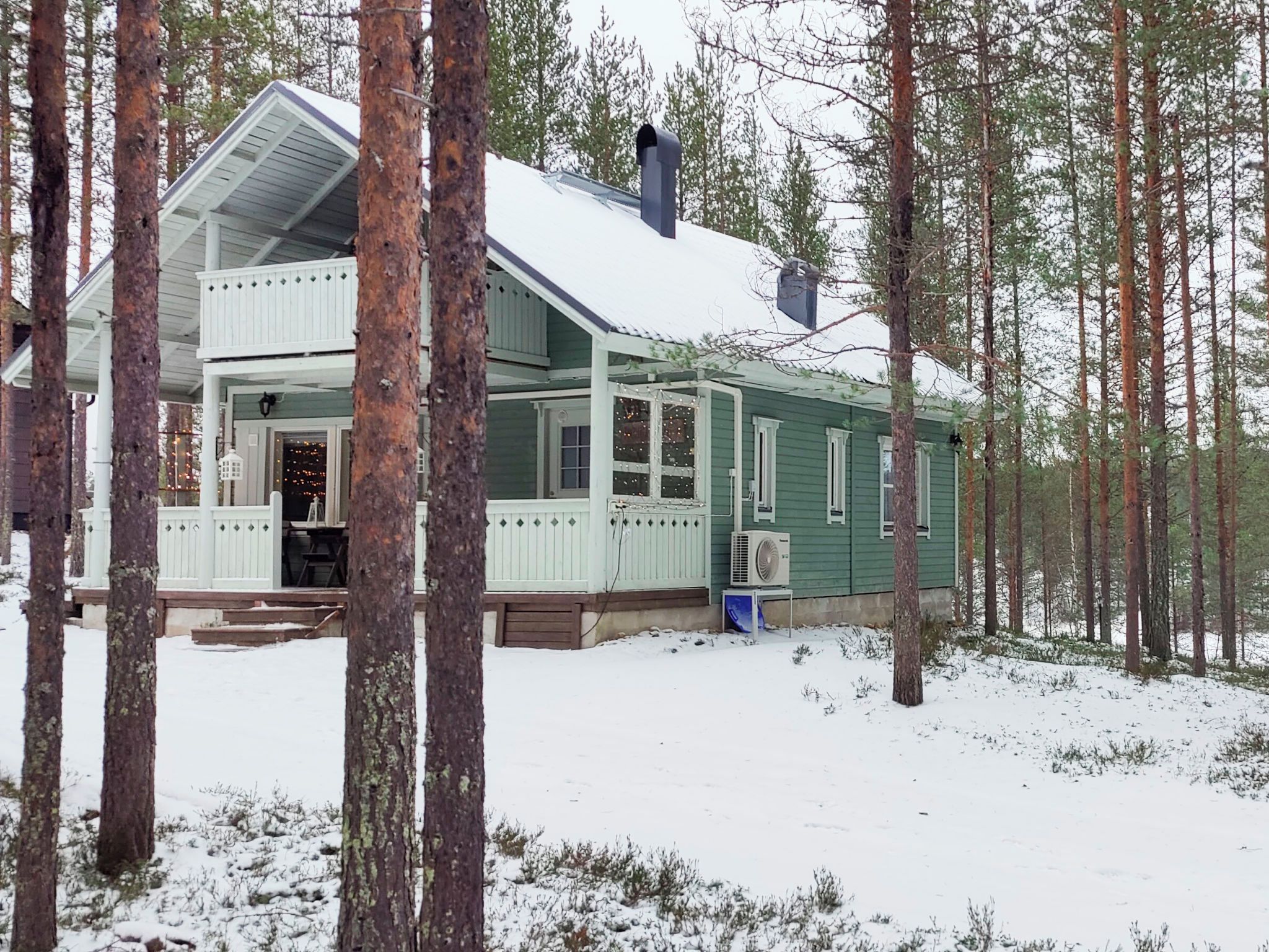A two-story house with a porch, surrounded by tall pine trees and covered in snow.