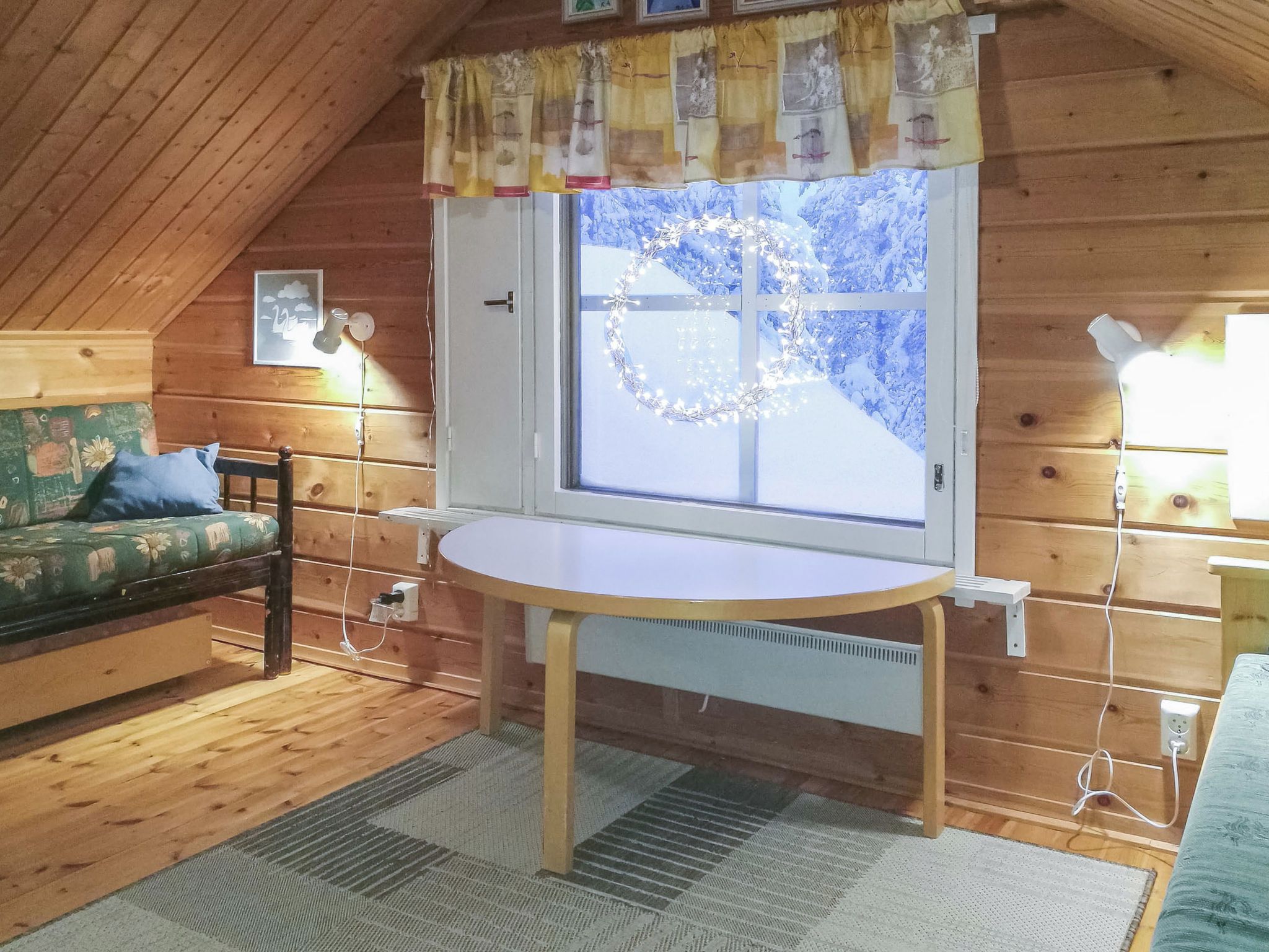 Een gezellige kamer met een houten plafond, een ronde tafel, een raam met een krans, een bank en een lamp.