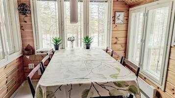 A wooden cabin with a dining table, chairs, potted plants, and a view of trees through large windows.