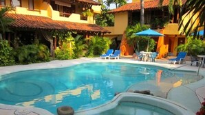 Una piscina con hidromasaje, rodeada de sillas de descanso y sombrillas, con un edificio al fondo.