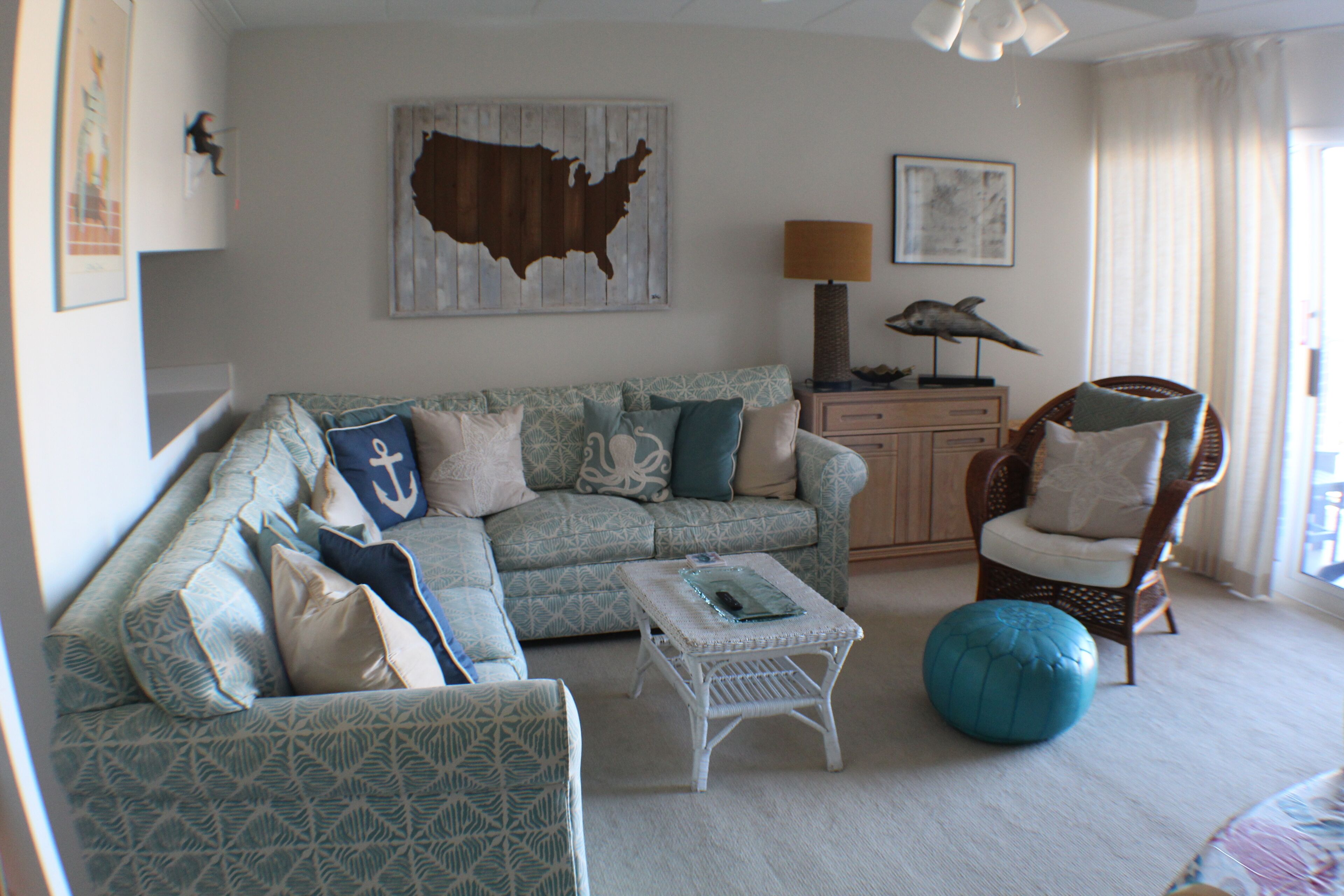 A living room with a patterned sofa, a coffee table, a blue ottoman, a wicker chair, and a map on the wall.