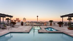 A rooftop pool with lounge chairs and potted plants, overlooking a city skyline at sunset.