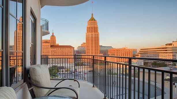 A balcony with a glass table and two chairs, overlooking a cityscape with prominent buildings and a clear sky.