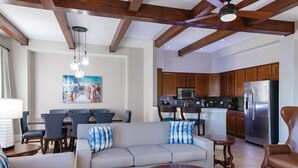 A modern living room with a sofa, coffee table, and dining area. It includes a kitchen with stainless steel appliances and wooden cabinets.