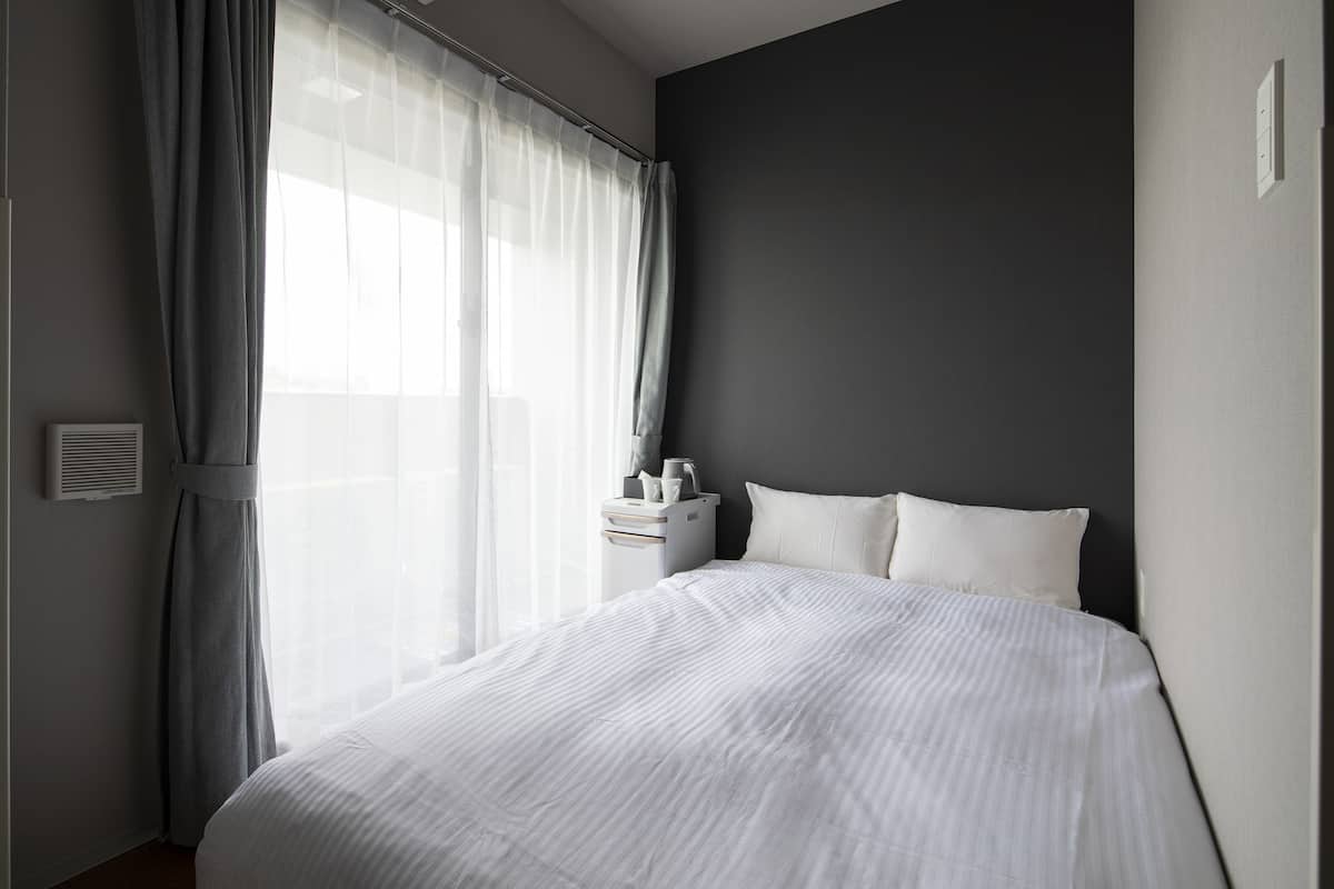 Una habitación de hotel moderna con una cama grande, una mesita de noche con lámpara y cortinas translúcidas en la ventana.