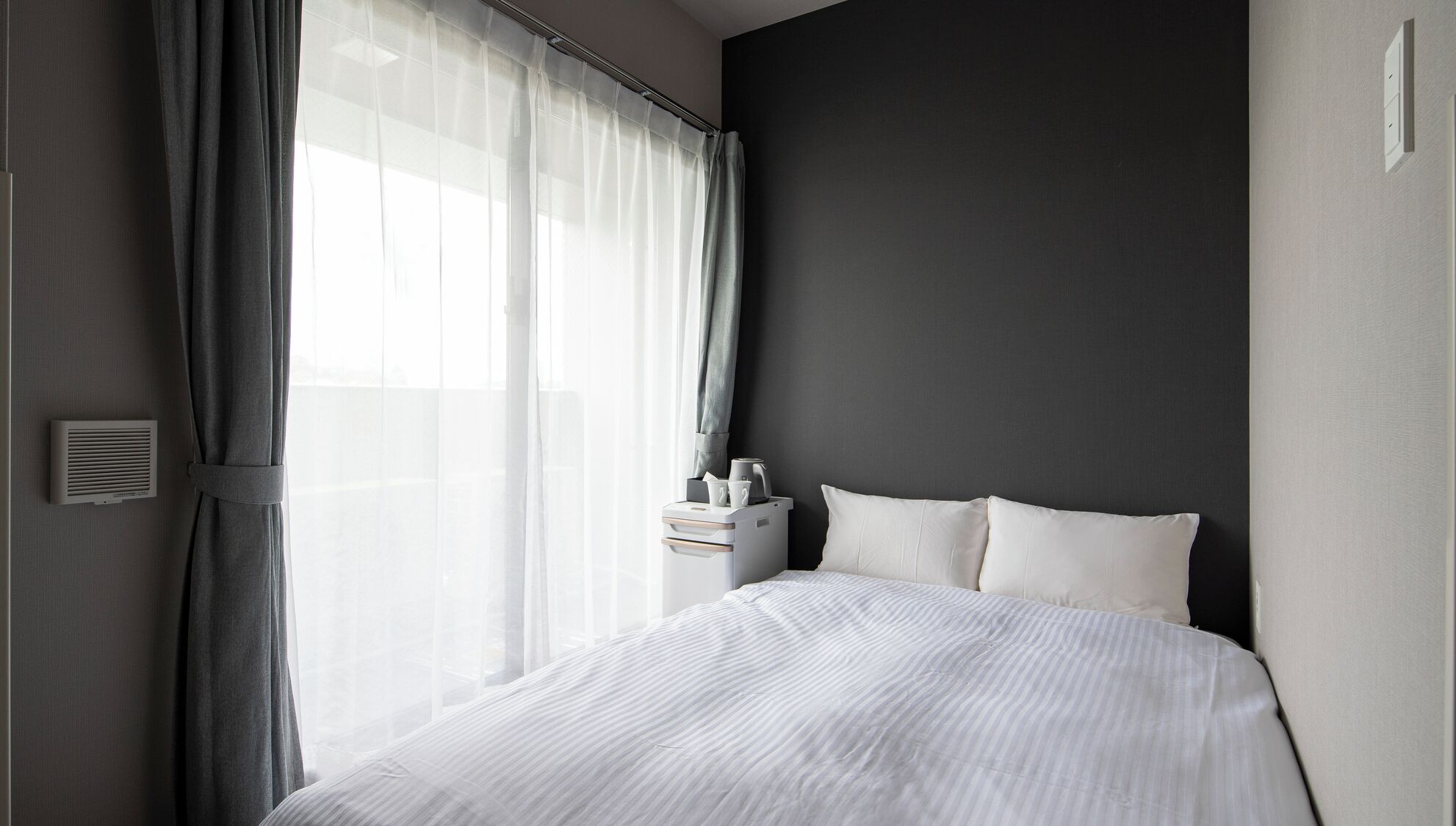 Une chambre d’hôtel moderne avec un grand lit, une table de chevet avec une lampe et une fenêtre ornée de rideaux légers.