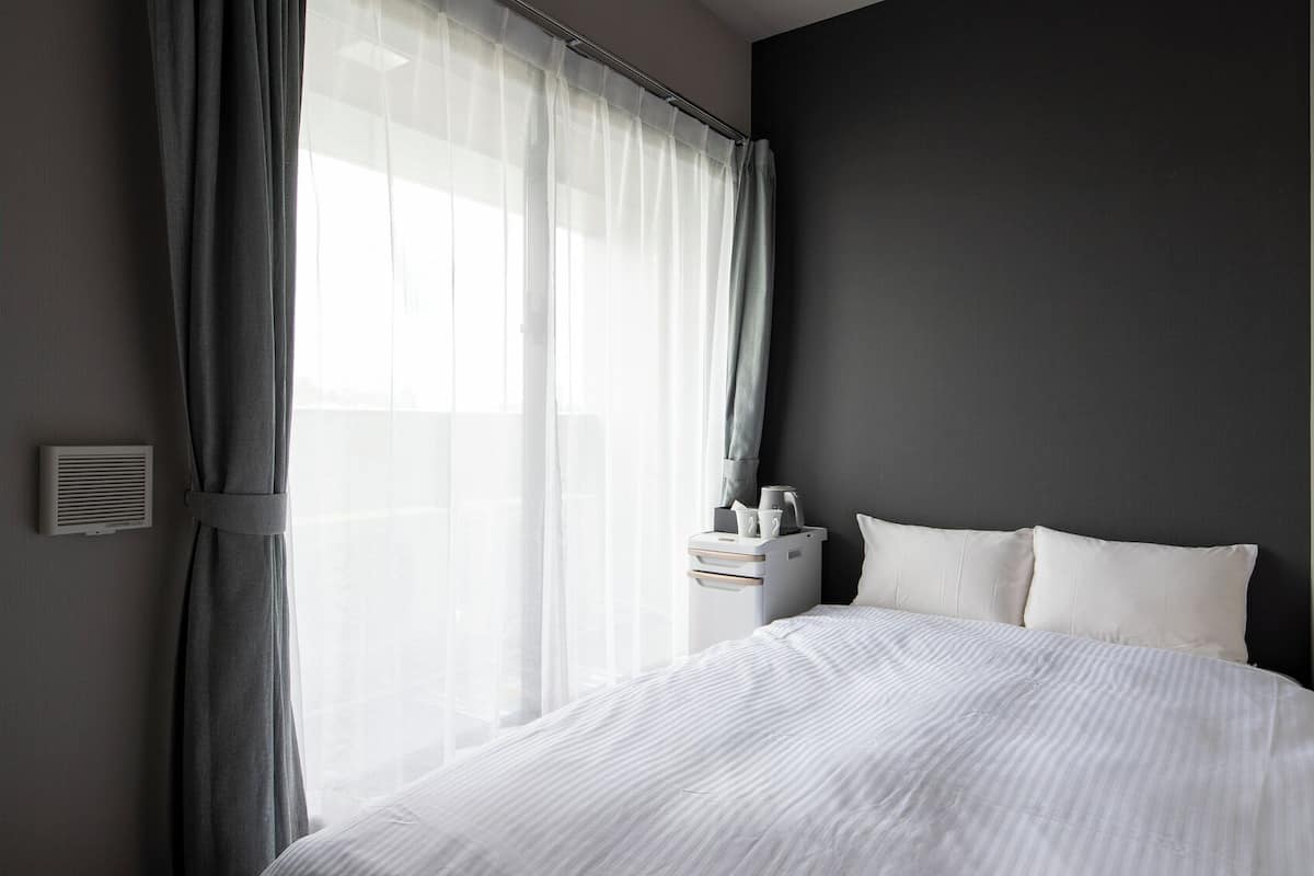 Una habitación de hotel moderna con una cama grande, una mesita de noche con lámpara y una ventana con cortinas translúcidas.