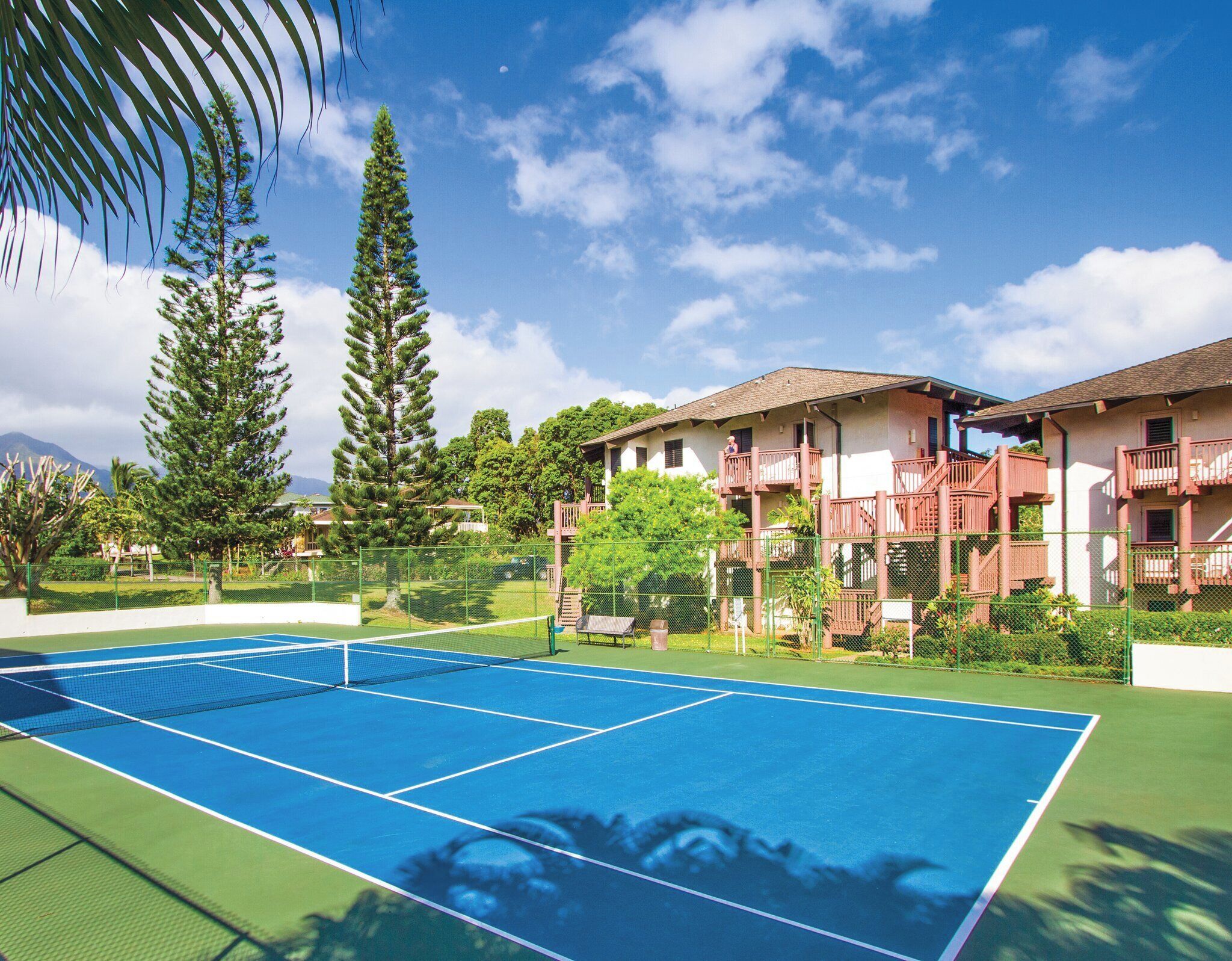 A tennis court with a net, surrounded by a fence, and adjacent to a building with balconies.