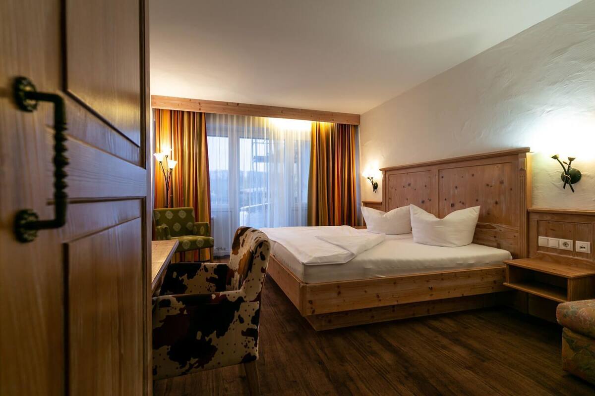 Ein Hotelzimmer mit einem hölzernen Bett, einem Schreibtisch, einem Sessel und Blick auf die Stadt durch große Fenster.