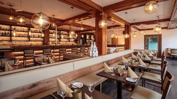A restaurant with wooden beams, round hanging lights, and a bar area with bottles. Tables are set with glasses and napkins.