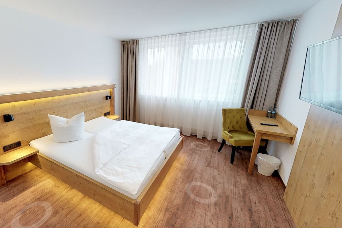 Ein Hotelzimmer mit einem Bett mit Holz-Kopfteil, einem Schreibtisch, einem Sessel und einem großen Fenster mit Vorhängen.
