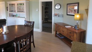 A dining area with a wooden table and chairs, a kitchen with white cabinets, a clock, a framed picture, and a lamp.
