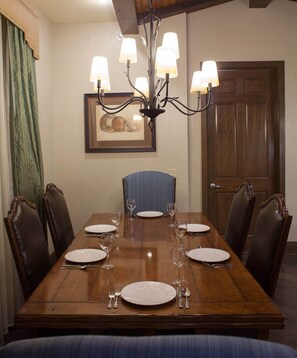 Un comedor con una mesa de madera dispuesta para seis comensales, rodeada por seis sillas. Un candelabro cuelga del techo, y hay un cuadro enmarcado en la pared.