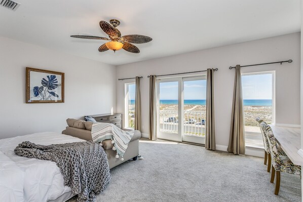 A bedroom with a bed, a ceiling fan, a large window with curtains, a view of the beach, and a dining area with a table and chairs.