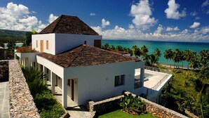 A modern two-story house with a tiled roof, white walls, and a stone pathway leading to the entrance. It overlooks a turquoise ocean with palm trees and a clear blue sky. - Luxury Contemporary Villa with Sea View & Private Pool – Las Terrenas STARLINK (Las Terrenas)