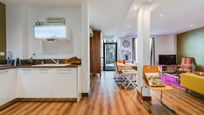 Una cucina moderna con mobili bianchi, un'isola in legno e una TV integrata. Si intravede una zona pranzo con tavolo e sedie, e un divano giallo si trova nel soggiorno.