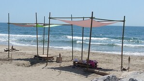 Una spiaggia con una struttura in legno che offre ombra, una piccola panchina in legno e un banchetto improvvisato con della merce.