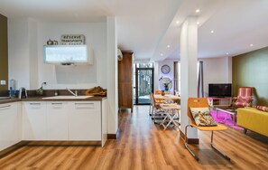A modern kitchen with white cabinets, a wooden island, and a built-in TV. A dining area with a table and chairs is visible, and a yellow sofa is in the living room.