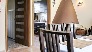 Uma sala de jantar com mesa e cadeiras de madeira, uma cozinha ao fundo e um candeeiro fixado na parede.