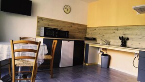 Een kleine keuken met een magnetron, koelkast en spoelbak. Er staat een houten stoel bij een tafel met een kantelaar. Een klok hangt aan de muur.