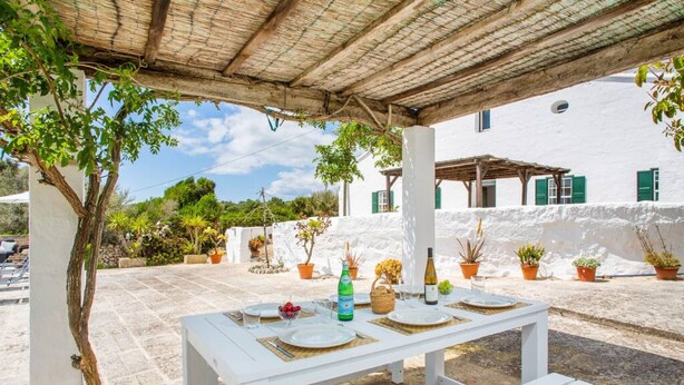 An outdoor dining area with a white table set for four, surrounded by potted plants and a white building with green shutters in the background. - Binicalaf · ❤️Finca Binicalaf Vell (Maó)