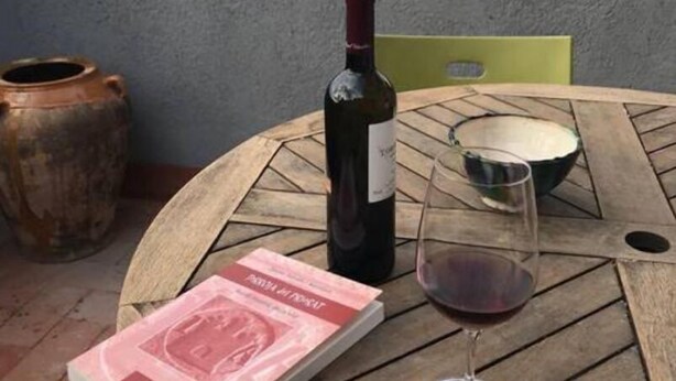 Ein Tisch mit einer Weinflasche, einem Weinglas und einem Buch auf einer Terrasse mit Blick auf Berge und Gebäude.