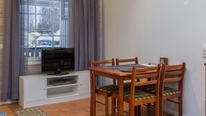 Un petit coin repas avec une table et des chaises en bois, un téléviseur posé sur un meuble blanc et une fenêtre avec des rideaux.