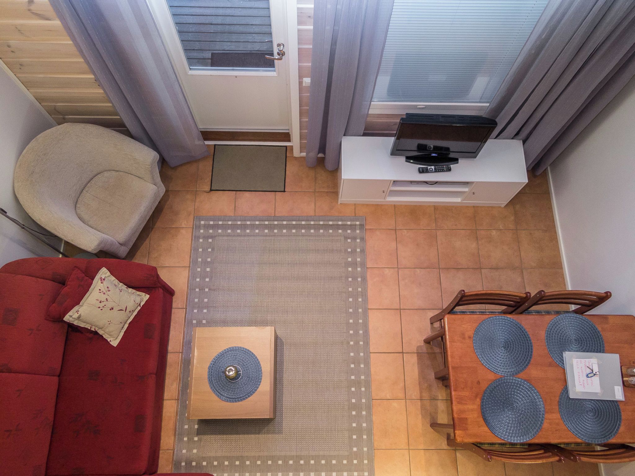 A compact living space with a red sofa, a wooden coffee table, a small side table, a television on a white cabinet, and a dining table with blue placemats.