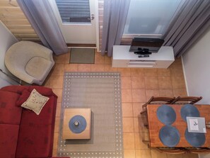 Ein gemütlicher Wohnbereich mit einem roten Sofa, einem Holztisch, einem kleinen Beistelltisch, einem Fernseher auf einem weißen Schrank und einem Esstisch mit blauen Tischsets.