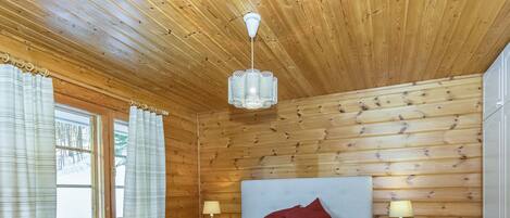 Un dormitorio de cabaña de madera con una cama, dos lámparas de noche, un ventanal con cortinas y una lámpara colgante.