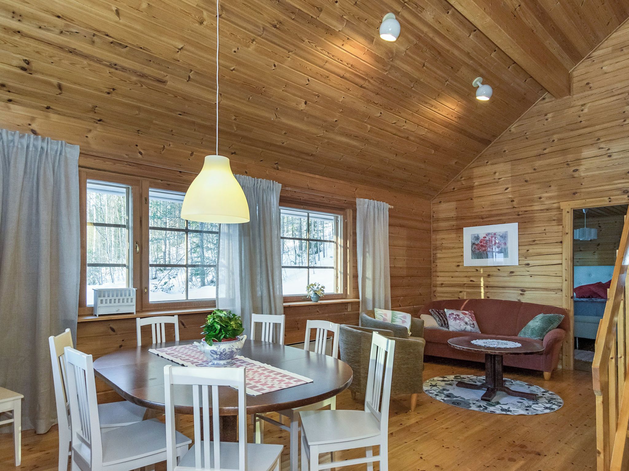 A wooden cabin interior with a dining table, chairs, a sofa, and a painting on the wall.