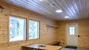 Interior de uma cabana de madeira, com uma mesa grande e bancos, duas janelas com vidro jateado e uma porta.