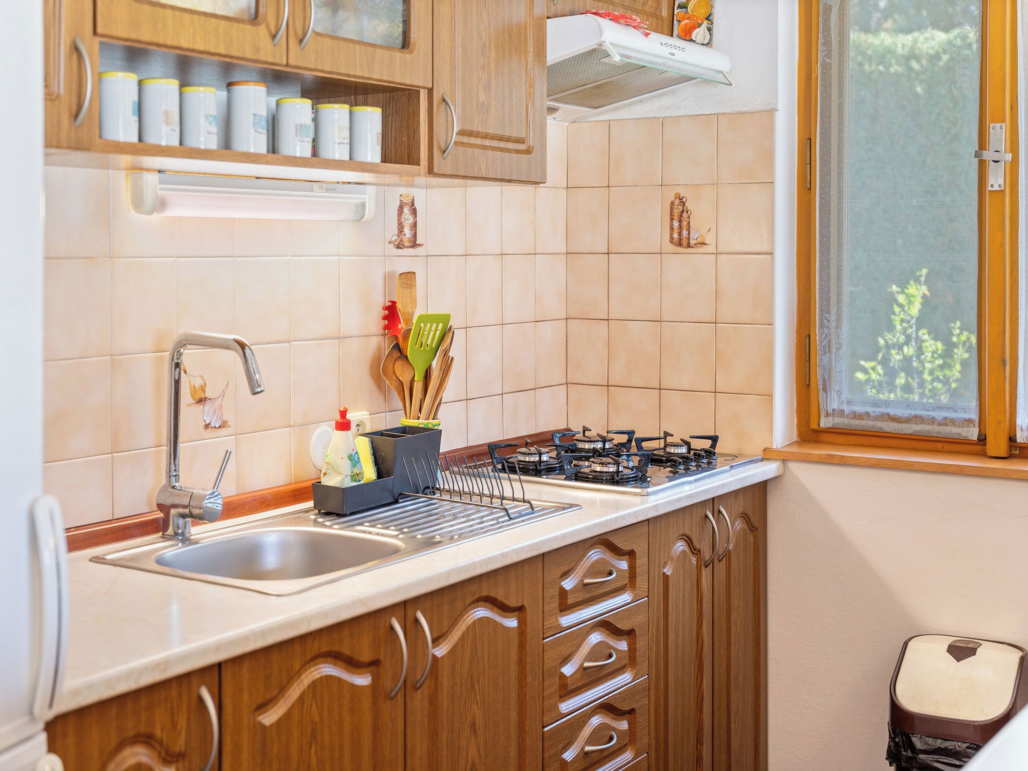 Una cucina con piano cottura a gas, lavello in acciaio inossidabile, mobili in legno e una finestra vista verde.