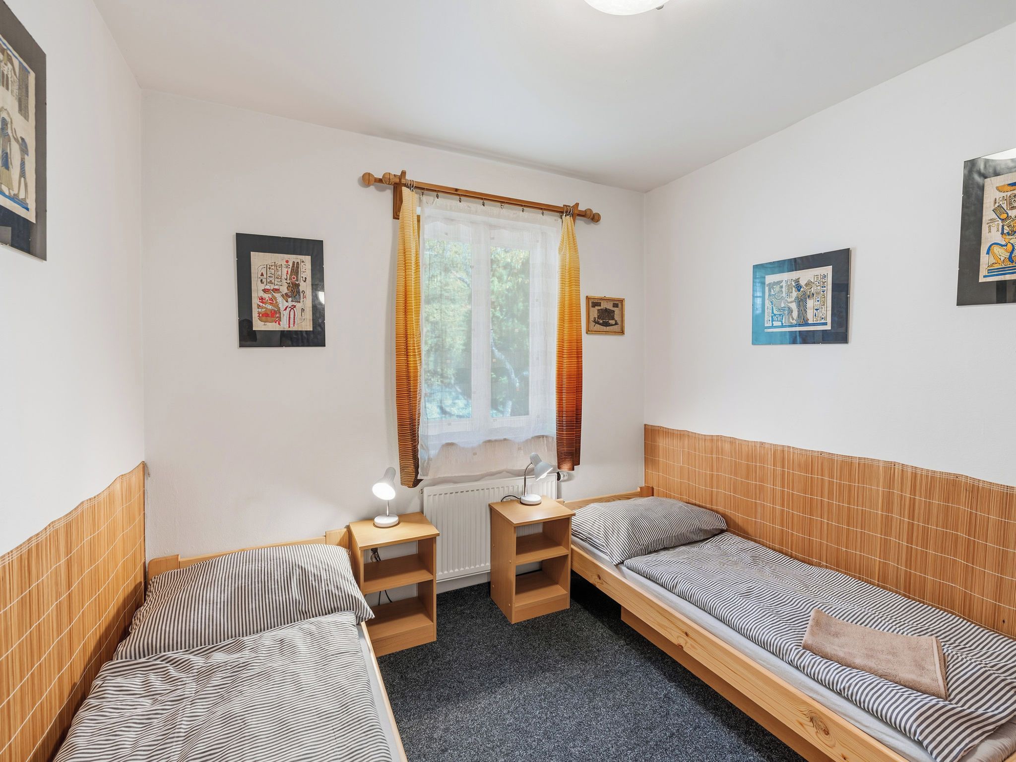 A small, cozy room with two single beds, a window with curtains, a radiator, and framed pictures on the walls.