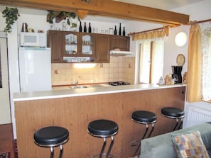 Eine Küche mit einer zentralen Kochinsel, Holzschränken, einer Mikrowelle und einem Spülbecken. Es gibt drei Barhocker und ein Fenster mit Vorhängen.