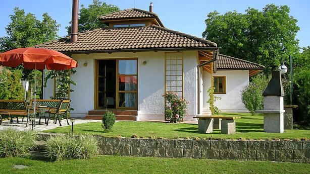 Una casa de dos pisos con techo de tejas, chimenea y jardín con banca y sombrilla.
