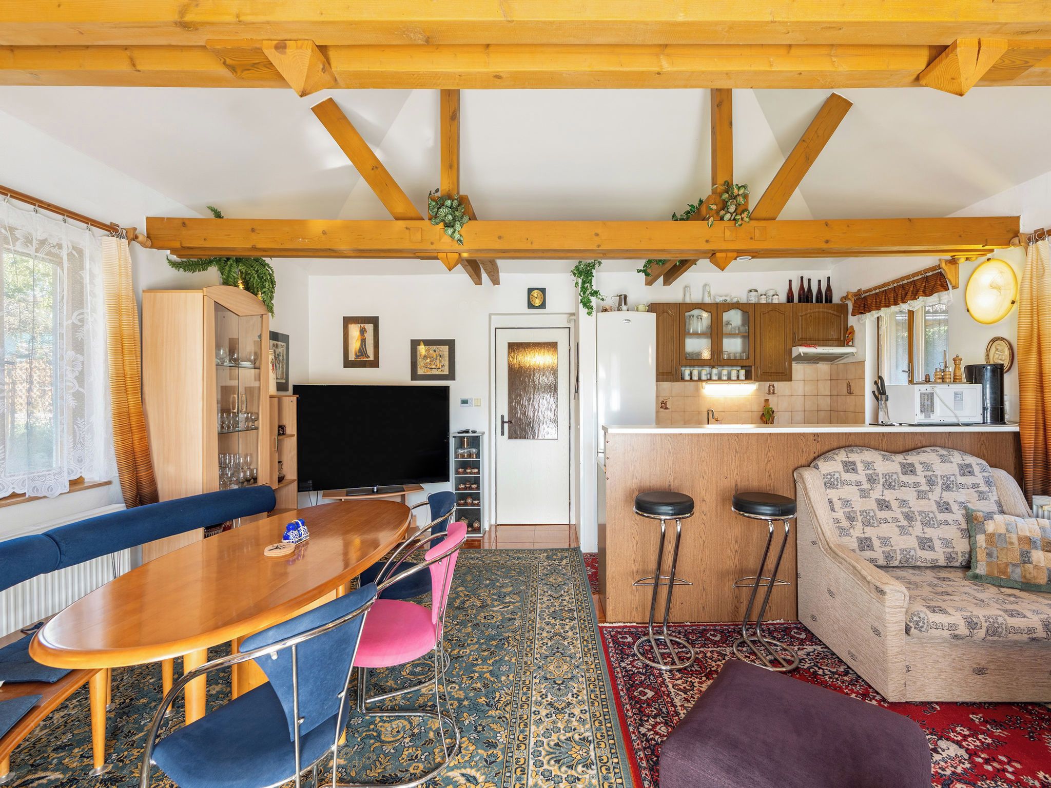 Un salon chaleureux avec un plafond en bois, un coin repas avec une table en bois et des chaises bleues, un canapé agrémenté de coussins à motifs et un coin cuisine avec des meubles en bois et un réfrigérateur.