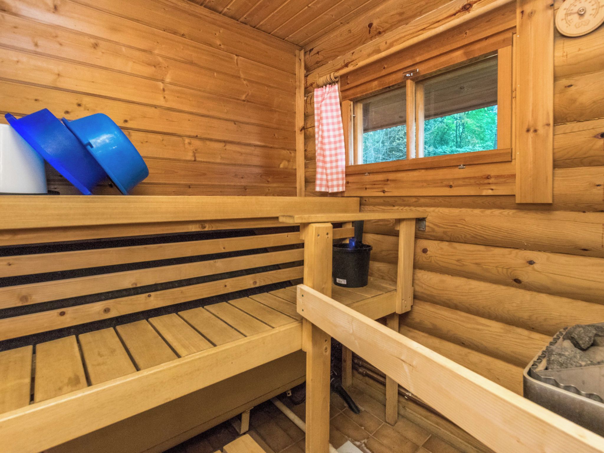 A wooden cabin interior with a bench, a window with a checkered curtain, and a black container on a shelf.