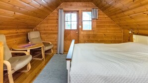 A cozy attic bedroom with a wooden sloped ceiling, a single bed, two chairs, a small table, and a window with curtains.