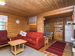 A cozy log cabin interior with a red sofa, wooden walls, a kitchen area, and a dining table.