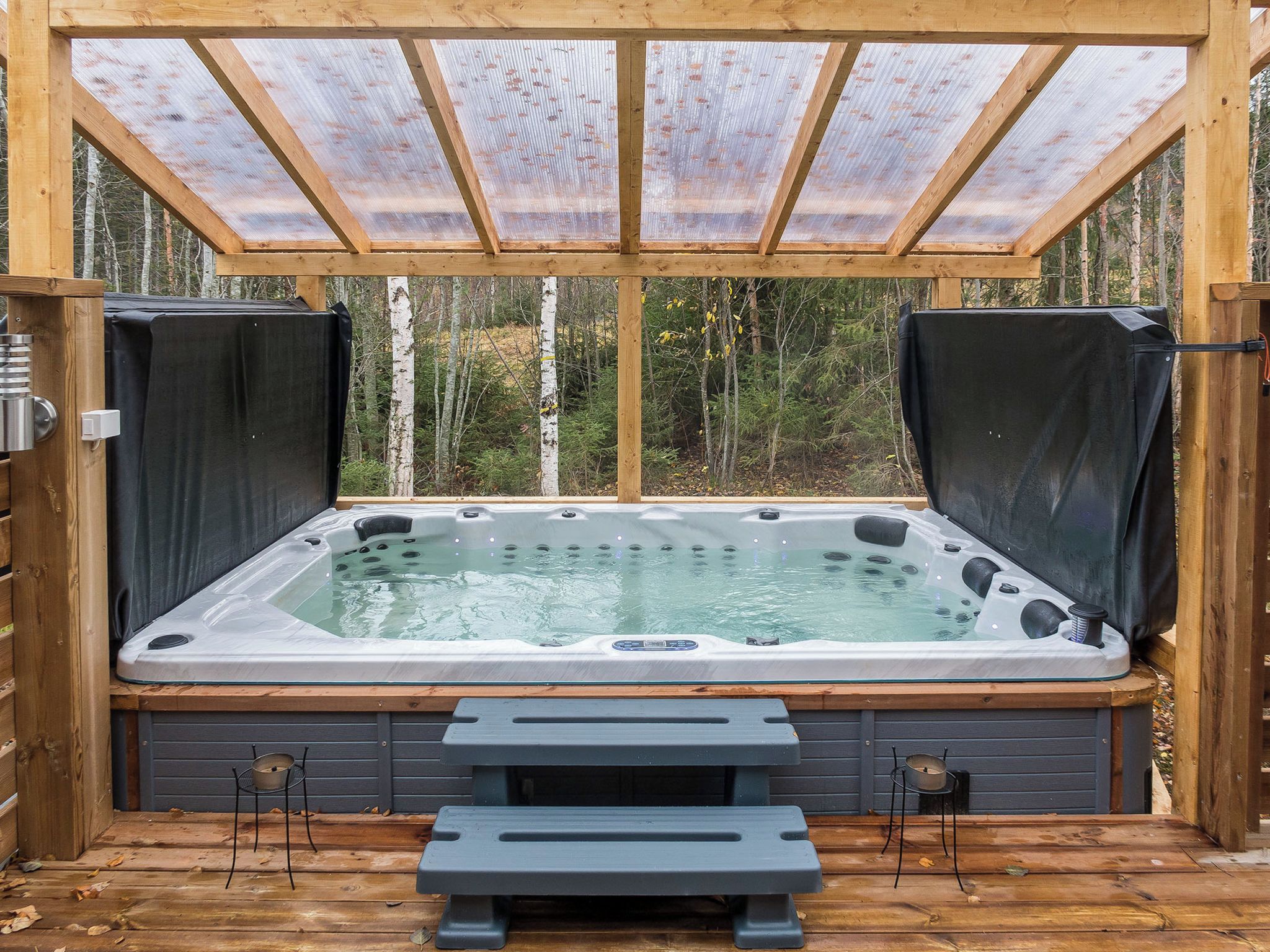 Un bain à remous doté d’une terrasse en bois, entouré d’une structure en bois surmontée d’un toit de verre, situé dans une zone boisée.